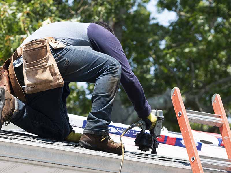 Workers using a nail gun to install GAF shingles on a residential roof (some packs of GAF shingles can be seen in the background)
