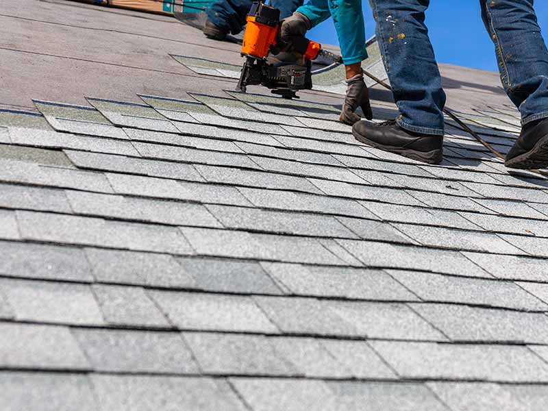Worker installing gray shingles on a residential roof with a nail gun